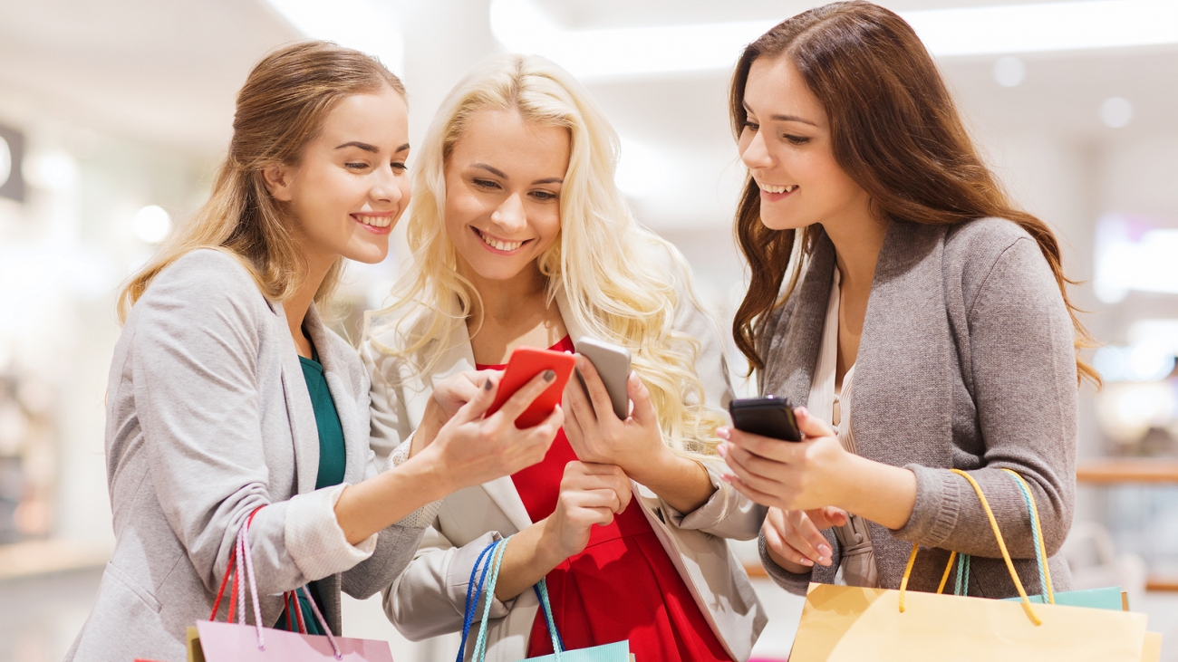 sale, consumerism, technology and people concept - happy young women with smartphones and shopping bags in mall