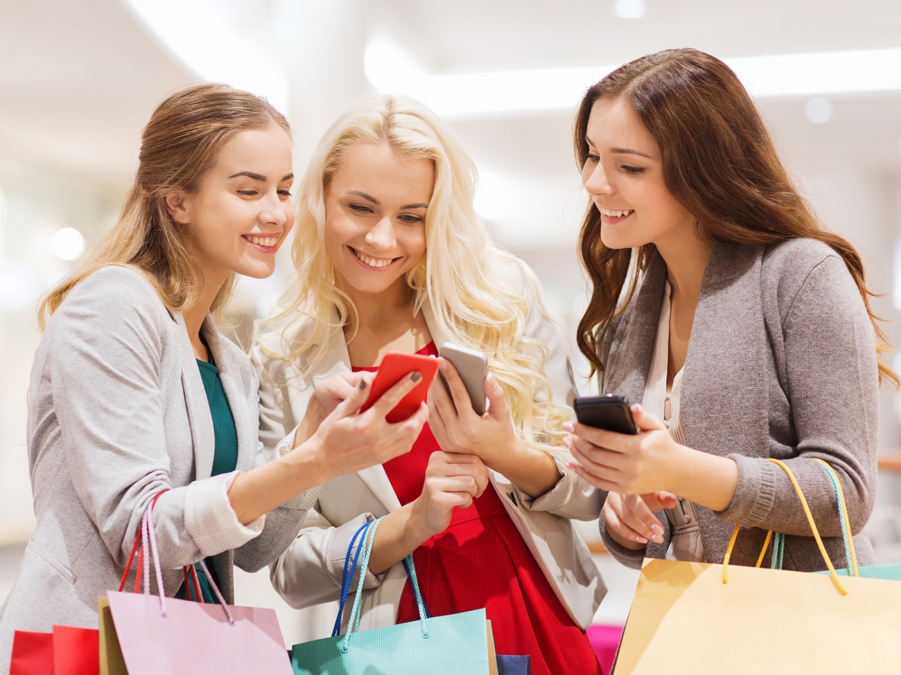 sale, consumerism, technology and people concept - happy young women with smartphones and shopping bags in mall