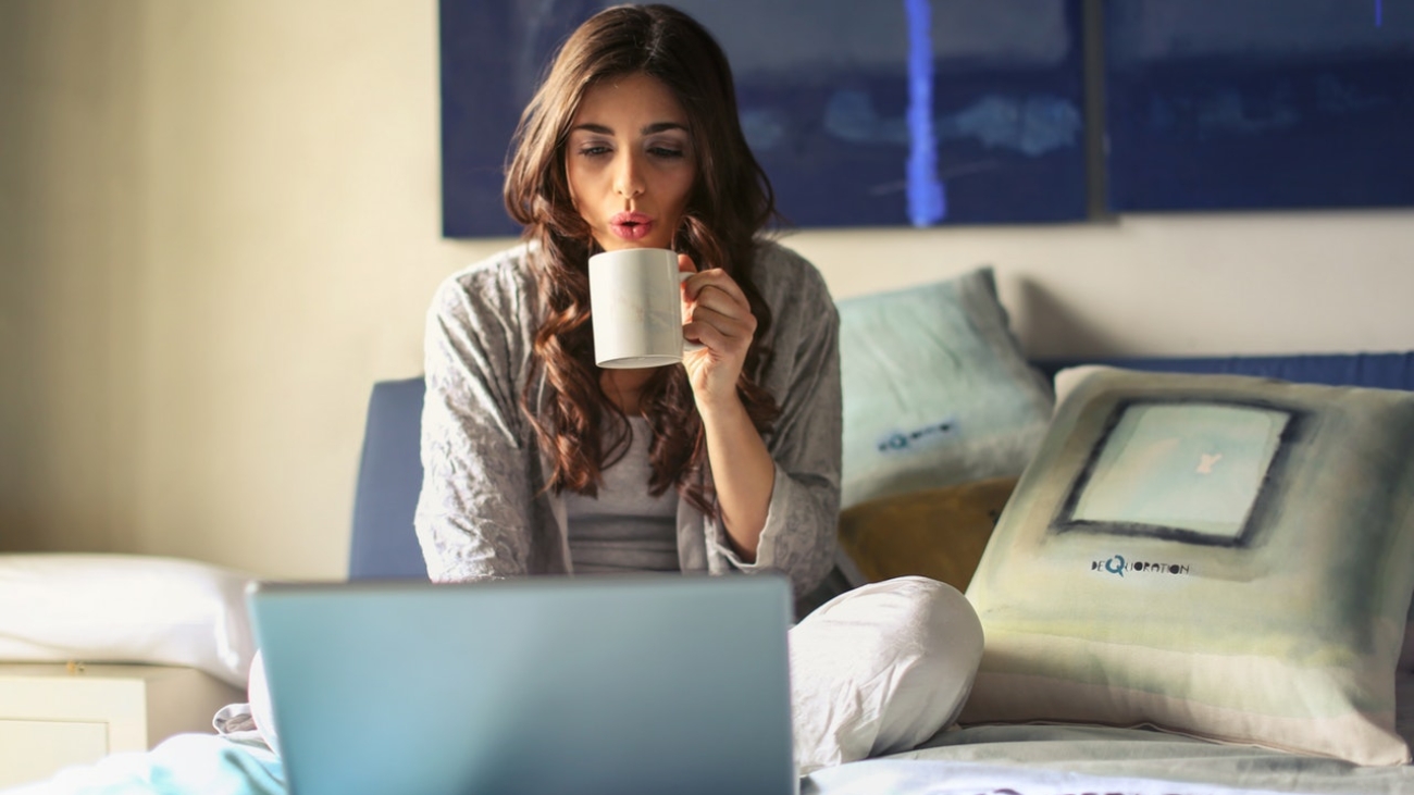 woman-in-grey-jacket-sits-on-bed-uses-grey-laptop-935743